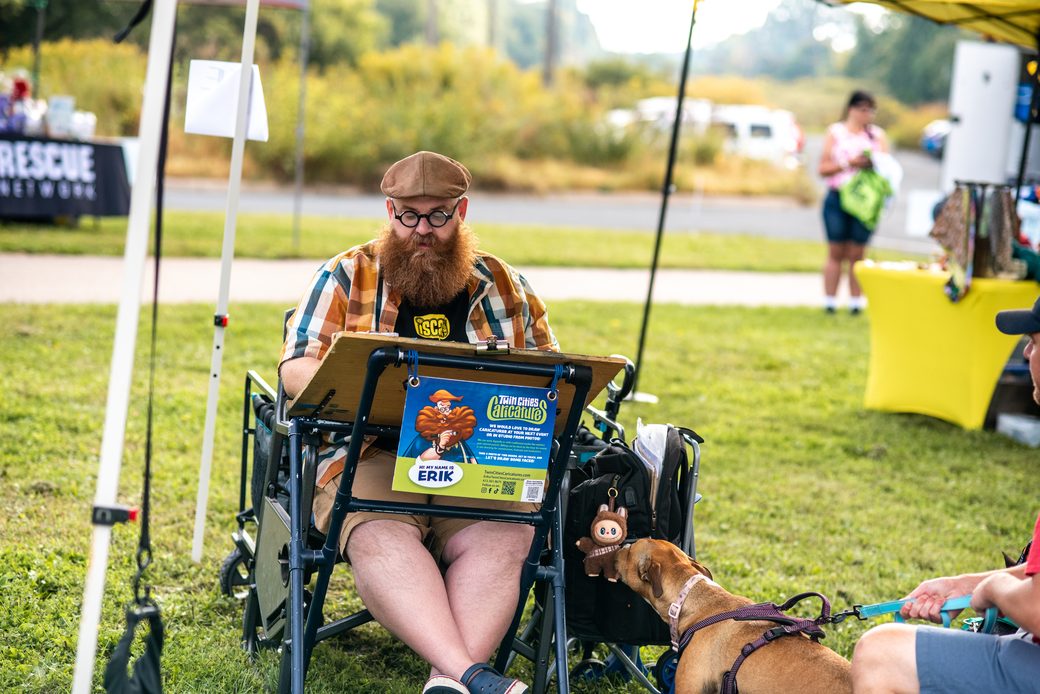 A caricature artist from Twin Cities Caricatures drawing under a tent while a curious dog sniffs his bag